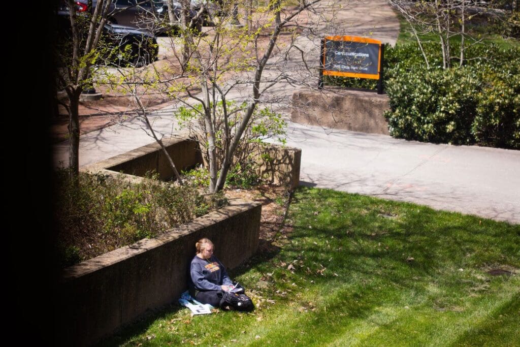 A student sits on the grass in front of the College of Communication and Information Building.