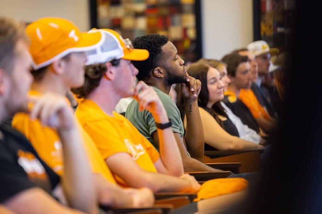 Students in orange gear listen to a sports communication guest speaker.